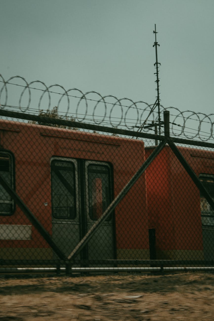An urban train passing behind a barbed wire fence during the day.