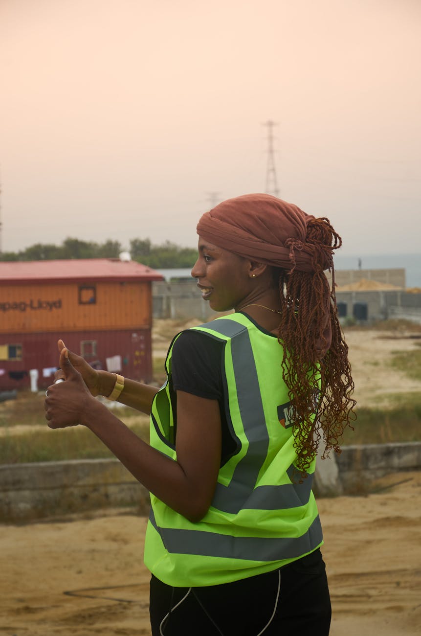 Confident female construction worker in safety vest giving thumbs up at a building site.