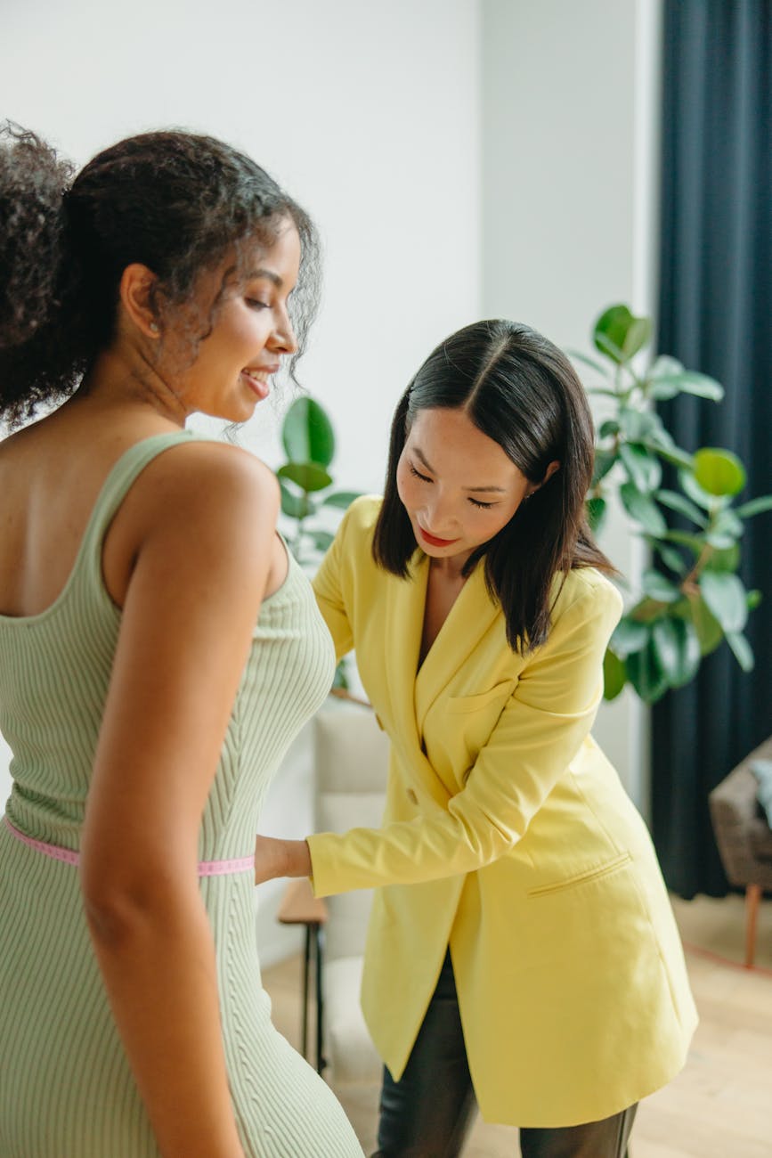 Two women measuring waistline indoors, focusing on fitness and fashion.