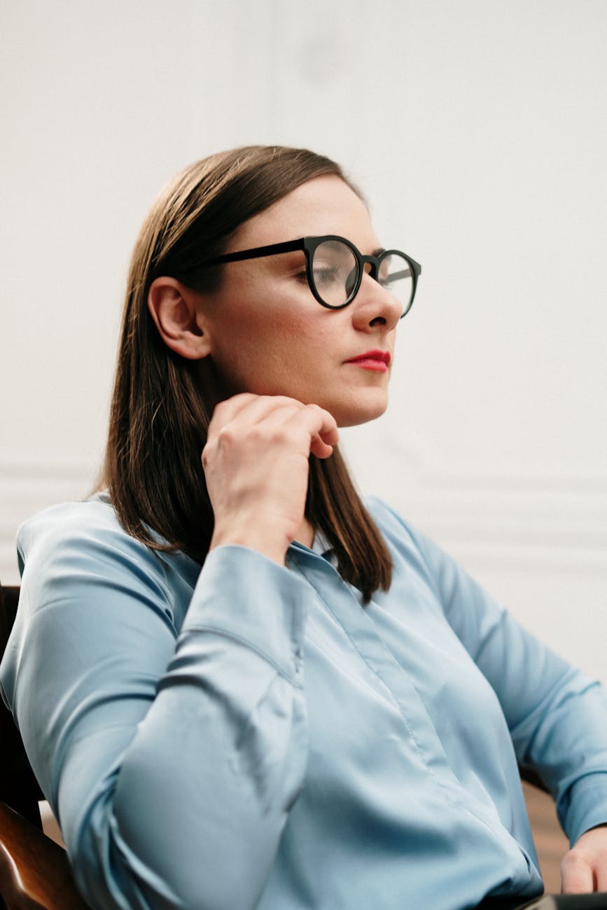 A thoughtful woman in glasses and a blue shirt sits indoors, exuding confidence.