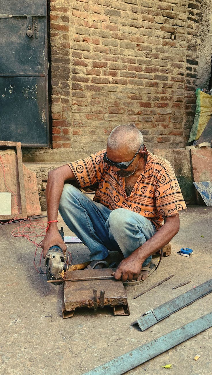 Elderly artisan using power tools for metalwork outside a rustic building in India.