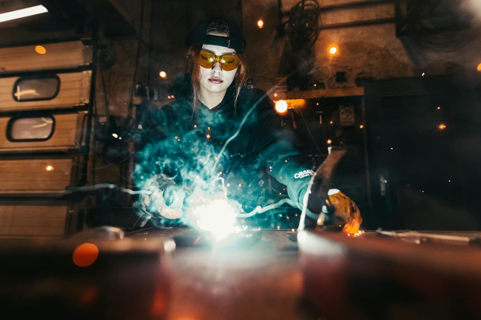 A welder wearing safety goggles working in a workshop filled with sparks and smoke.