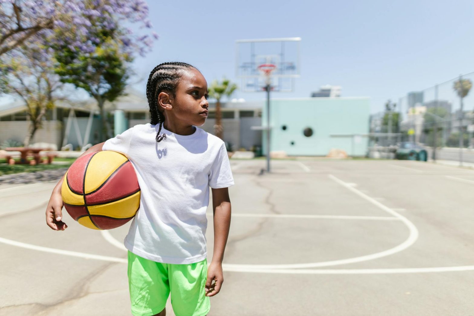 Young boy with braided hair holding basketball under clear sky on an outdoor court.