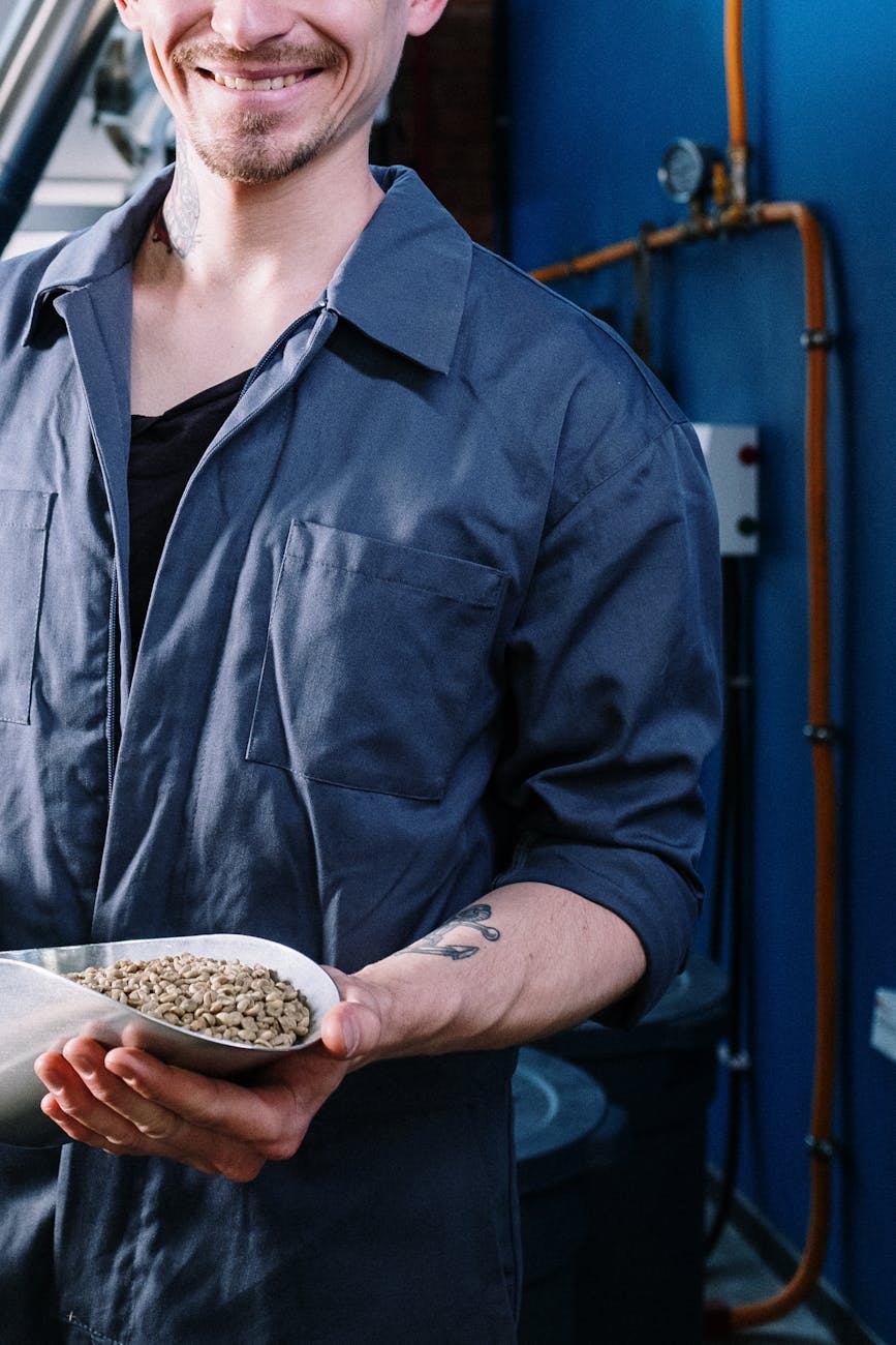 A man in a factory uniform smiles while holding a scoop of unroasted coffee beans indoors.