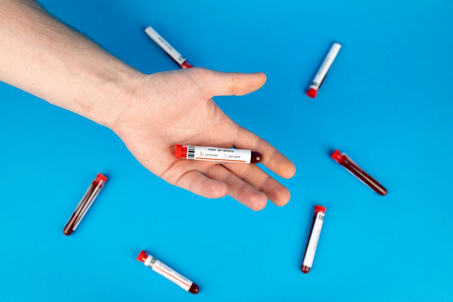 Close-up of a hand holding a blood sample vial on blue background, representing COVID-19 testing.