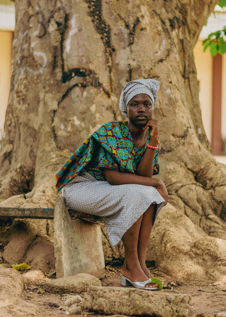 Thoughtful woman seated by a tree, wearing colorful traditional African clothing.