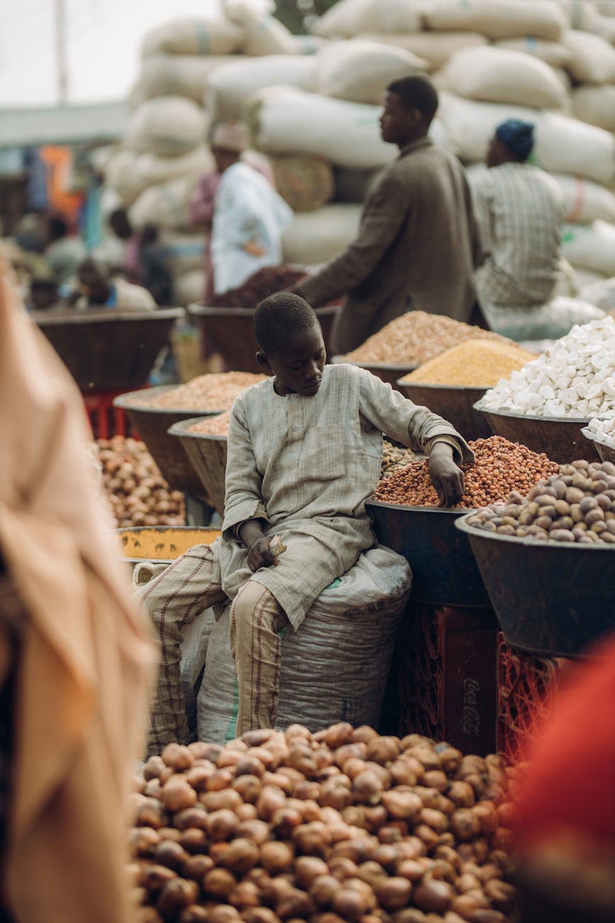 A young boy sits amidst a bustling market surrounded by sacks and bowls of grains and nuts.