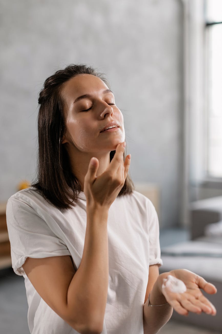 Young woman applying skincare cream to her face in a bright indoor setting.