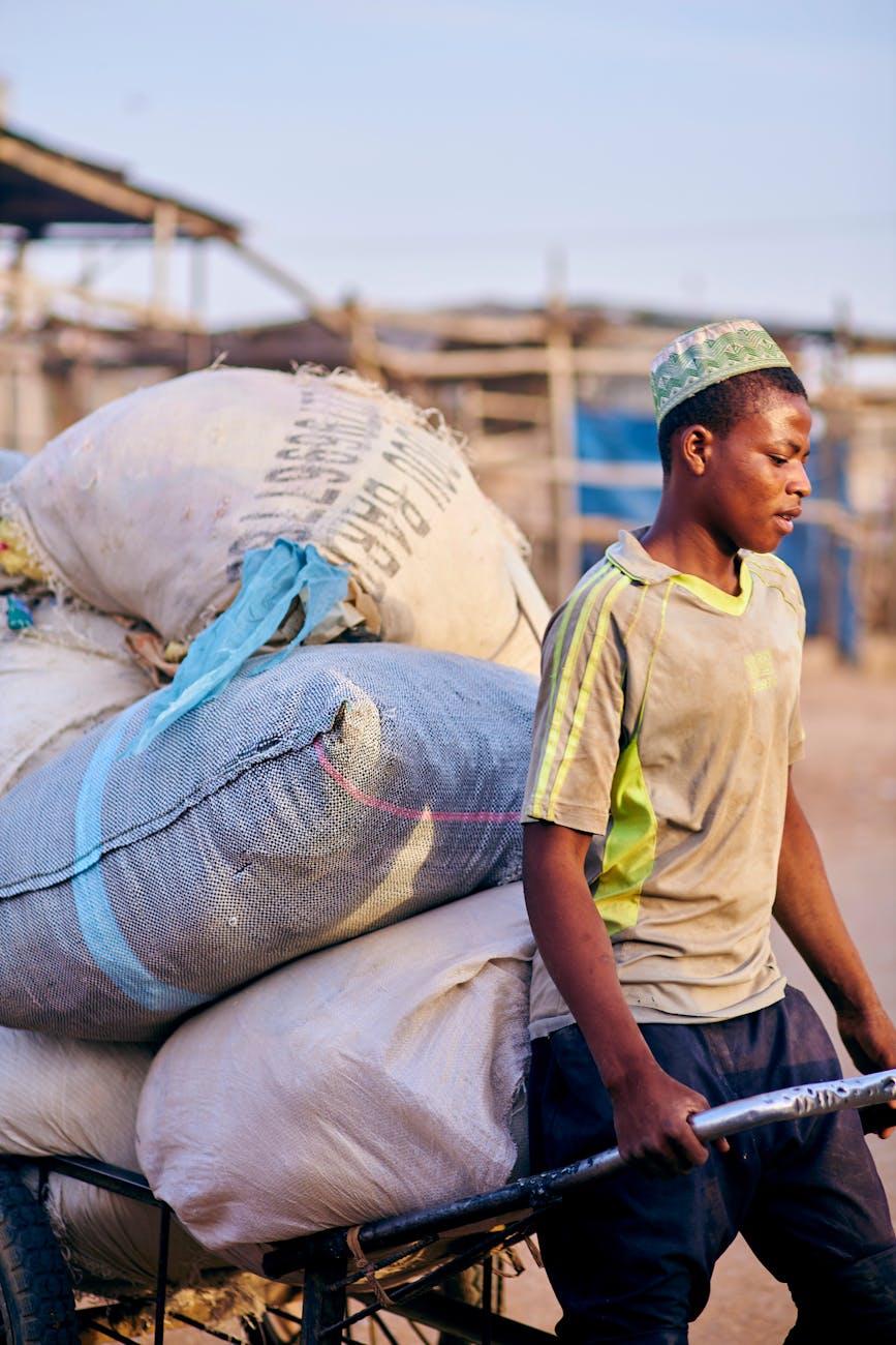 Dedicated young man moving large goods on wheelbarrow in a bustling marketplace at sunset.