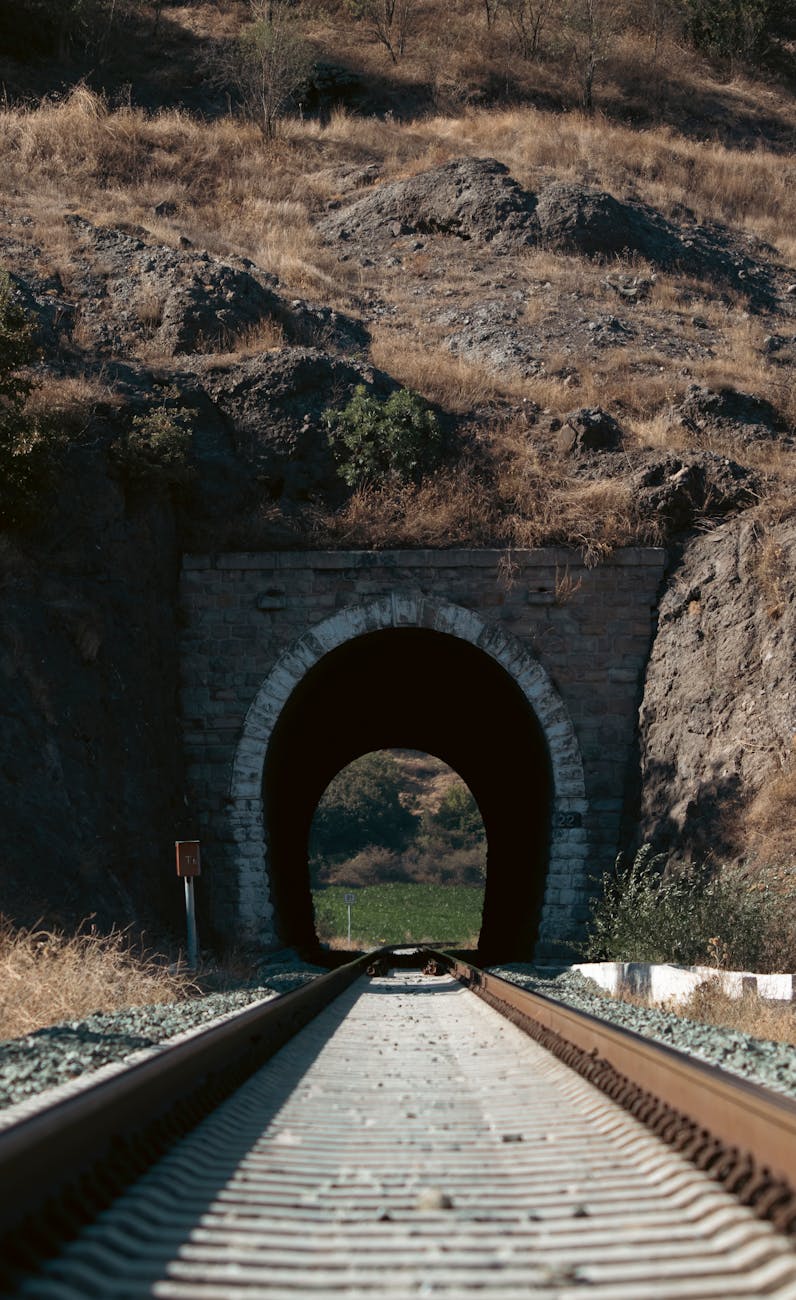 A railway track leading into a rustic mountain tunnel in a dry, rocky landscape.